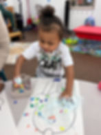 A young child in a graphic t-shirt is sitting at a table making a craft project.